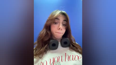 Media: Video of a young woman with long, wavy brown hair, wearing a white shirt with red \"You Have\" text, large black headphones, and a neutral expression against a blue background.