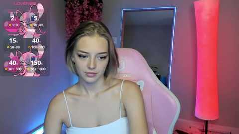 Media: A video of a young woman with light skin and straight brown hair, wearing a white spaghetti-strap top, seated in a pink gaming chair in a room with a pink light.