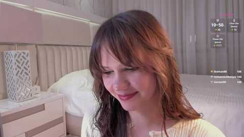 Media: Video of a smiling, fair-skinned woman with wavy brown hair, wearing a white sweater, in a modern bedroom with beige headboard, white bed, and patterned lamp.