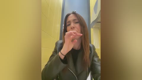 Media: Video of a woman in a yellow-tiled restroom, wearing a black leather jacket, holding a toilet brush in her mouth, appearing annoyed.