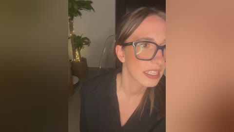 Media: Video of a woman with fair skin, glasses, and straight brown hair, smiling while looking to the right, standing in a dimly lit room with a potted plant on the left.