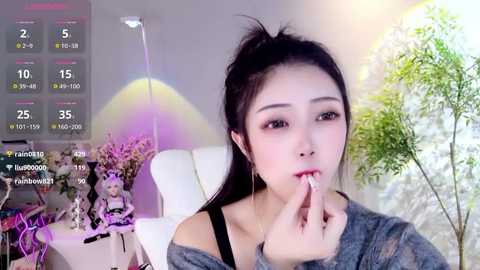 Media: Video of an East Asian woman with long black hair, wearing a grey sweater, biting her finger, in a bright, modern room with a white bed, green plants, and a purple lamp.