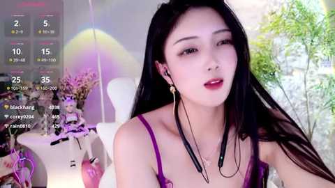 Media: Video of a young East Asian woman with long black hair, wearing a purple spaghetti-strap top, black dangling earrings, and red lipstick, sitting indoors with a colorful background.