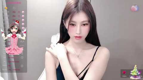 Media: Video of a young East Asian woman with pale skin, long black hair, and delicate features, wearing a black strapless top, captured in a selfie-style video. Background includes a gray wall and a video screen displaying a cartoon character.