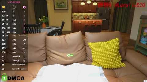 Media: Video of a cozy living room with a beige sofa, yellow polka-dot pillow, and a white cloth on the floor. Background features a modern kitchen with wooden cabinets, pendant lights, and a dining area with a table and chairs.