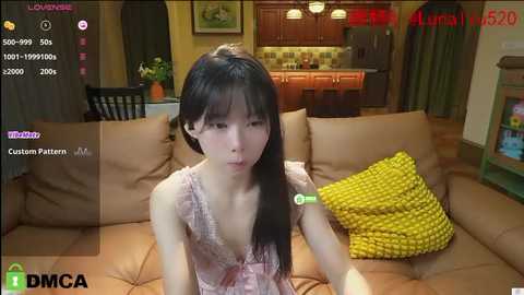 Media: A video of a young Asian woman with long black hair, wearing a pink lace dress, sitting on a beige couch in a cozy living room.