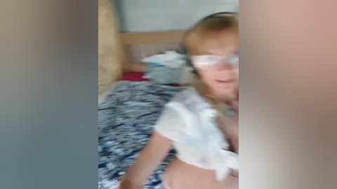 Media: Video of a young blonde woman in glasses, wearing a white crop top, lying on a bed with blue patterned sheets.