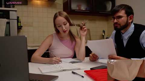 Media: Video of a young Caucasian woman with blonde hair in a pink sleeveless top, writing at a desk, next to a bearded man with glasses, holding papers in a modern kitchen.