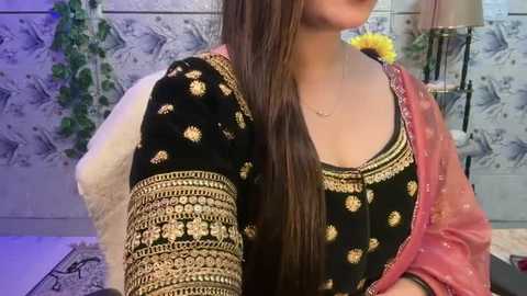 Media: Video of a woman with long, straight brown hair, wearing a traditional black and gold embroidered Punjabi suit with a red dupatta, against a floral wallpaper background.