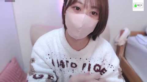 Media: A video of an Asian woman with short brown hair and fair skin, wearing a white sweater with \"I'm sorry\" in red, a pink face mask, and a white mask. She is indoors, possibly in a bedroom, with a blurred background.