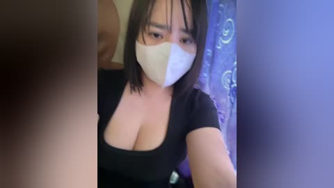 Media: Video of a young Asian woman with short black hair and a white surgical mask covering her nose and mouth, wearing a black top that reveals her large cleavage. Background features a blue curtain with white patterns.