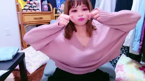 Media: Video of an East Asian woman with straight brown hair, wearing a pink sweater, playfully pulling the sweater up, in a cluttered bedroom with a wicker chair and various clothes.