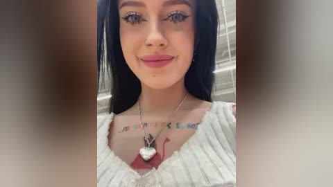 Media: Video of a young woman with fair skin, long black hair, and dark eye makeup. She wears a white, ribbed, off-shoulder top and a necklace with a heart pendant. Background is blurred.