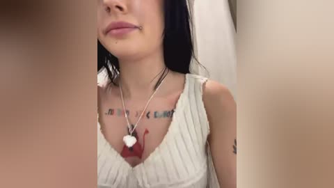 Media: Video of a young woman with light skin and long black hair, wearing a white sleeveless top with a plunging neckline, displaying a tattoo with text on her chest. She has a silver necklace with a heart pendant.