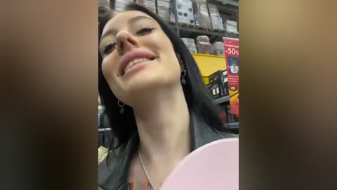 Media: Video of a smiling woman with long black hair in a store, wearing a black leather jacket, surrounded by shelves of products.