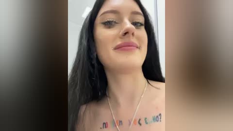 Media: Video of a young woman with long black hair, wearing a silver necklace, smiling with a septum piercing. She has tattoos on her shoulders.