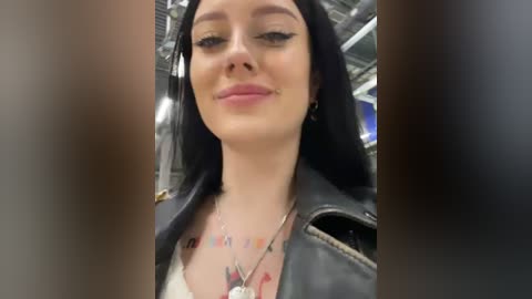 Media: A video of a young woman with long black hair, wearing a black leather jacket, silver necklace, and a white top. She has a faint smile and black eyeliner. The background shows an indoor setting with metallic structures.