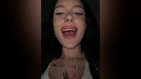 Media: Video of a young woman with pale skin, dark hair, and a silver septum piercing, wearing a white top, smiling widely with her mouth open.