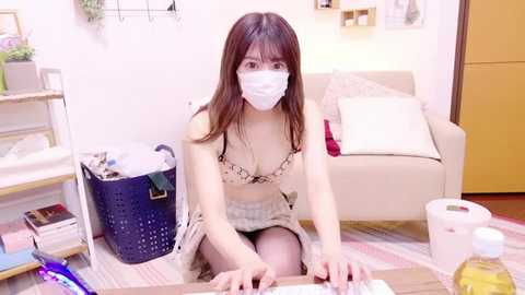 Media: Video of an Asian woman with light skin, long brown hair, wearing a white mask, and a beige bra with lace details, kneeling in a messy living room with a white couch, shelves, and a laundry basket.