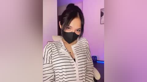 Media: Video of an East Asian woman with dark hair in a ponytail, wearing a black face mask, black and white striped shirt, and a white jacket, standing indoors with pastel-colored walls.