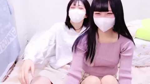 Media: Video of two young Asian women with long black hair, wearing white masks and pastel sweaters, sitting on a bed with white sheets, in a cozy indoor setting.