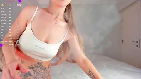 Media: Video of a light-skinned woman with long blonde hair, wearing a white sports bra, revealing a tattooed side. She is in a minimalistic, brightly lit bedroom with white walls and a bed.