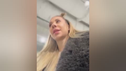 Media: Video of a blonde woman with a slight smile, wearing a gray fur coat, taken from a low angle, creating a distorted perspective.