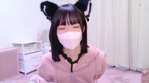 Media: Video of a young woman with straight black hair, wearing cat ears, a pink blouse, and a white mask in a pink room.