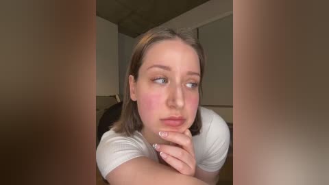 Media: A video of a young woman with light skin, shoulder-length brown hair, and pink blush on her cheeks, looking contemplative in a white top, indoors with beige walls and a wooden floor.