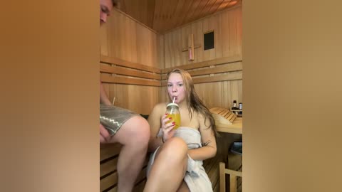 Media: Video of a blonde woman in a towel, drinking a juice in a wooden sauna, with another person partially visible in the background.