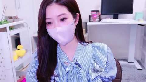 Media: Video of a young East Asian woman with long dark hair wearing a light blue striped blouse and a white face mask, seated in a modern office with white furniture and a computer.