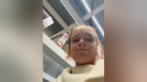 Media: A video of a young girl with fair skin and light brown hair, wearing a yellow sweater, standing in a supermarket aisle. The background features shelves with products and a metal ceiling grid.