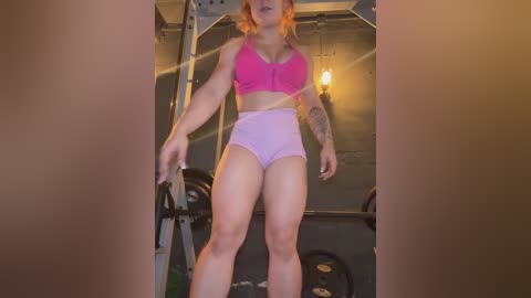 Media: Video of a fit, light-skinned woman with red hair, wearing a pink sports bra and high-waisted pink shorts, lifting weights in a dimly lit gym with metal beams and exposed pipes in the background.