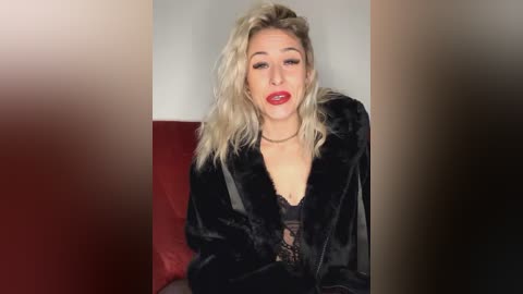 Media: Video of a blonde woman with wavy hair, wearing a black fur coat and a black lace top, sitting on a red cushioned chair against a plain beige wall. She has a confident expression with red lipstick.