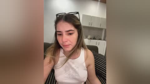 Media: Video of a young Caucasian woman with straight, light brown hair and a white tank top, sitting on a striped couch in a minimalist kitchen with white cabinets.