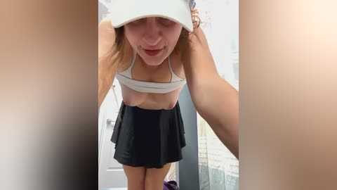 Media: A video of a young, fair-skinned woman with blonde hair, wearing a white visor and a black skirt, exposing her breasts in a bathroom with a glass shower door.