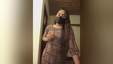 Media: A video of a South Asian woman with long dark hair, wearing a black face mask, a patterned brown dress, and standing in a narrow hallway with beige walls and wooden ceiling.