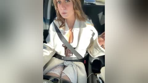 Media: Video of a woman with long blonde hair, wearing a white shirt with a brown tie, and light-colored pants, driving a car with a safety belt on.