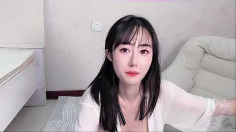 Media: A video of a young Asian woman with long black hair, wearing a white lace robe, sitting on a striped rug in a minimalist room with white walls and a white leather chair.