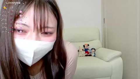 Media: Video of an Asian woman with long black hair, wearing a white mask, sitting on a white leather chair in a minimalist room with a stuffed Mickey Mouse toy.