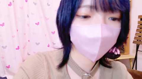 Media: Video of an Asian woman with short blue hair, wearing a light pink surgical mask, beige sweater, and a necklace, in a room with a pink heart-patterned curtain.