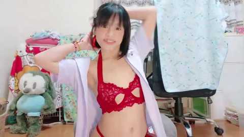 Media: Video of an Asian woman with short black hair and light skin, wearing a red lace bra and panties, posing in a bedroom with plush toys, clothing, and a sewing machine in the background.