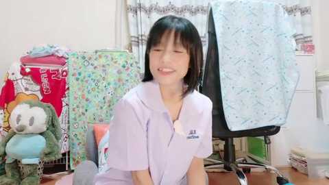 Media: Video of a young Asian woman with short black hair, wearing a light purple blouse, smiling while sitting on the floor in a cluttered room with colorful stuffed animals, fabric, and a sewing machine.