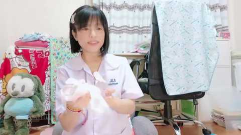 Media: A video of an Asian woman with black hair, wearing a white uniform, holding a white cloth in a cluttered, brightly lit room with toys and laundry.