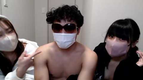 Media: Video of three East Asian individuals in a clinical setting. One man with curly hair, wearing a white mask, sits center. Two women, masked, flank him; one points, the other holds a cloth.