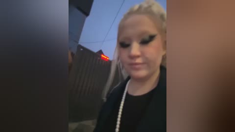 Media: Video of a blonde woman with heavy black eyeliner and a pearl necklace, standing outside near a dark fence with a red light.