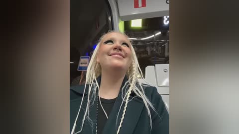 Media: A video of a young, smiling woman with long, braided blonde hair, wearing a dark jacket and black shirt, standing in an airplane cabin.