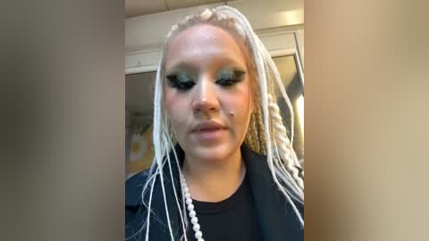 Media: Video of a young woman with light skin and platinum braids, wearing dramatic blue eyeshadow and a black jacket, standing in a dimly lit room with beige walls and a mirror.