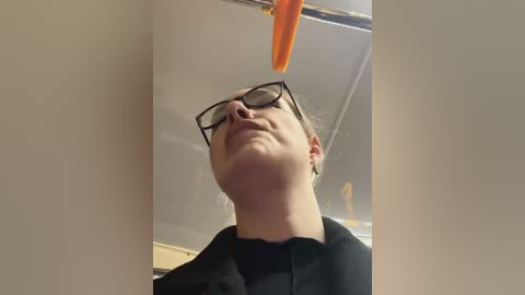 Media: Video of a young woman with glasses and light skin, wearing a black shirt, looking up at a dangling orange carrot in a cafeteria ceiling.