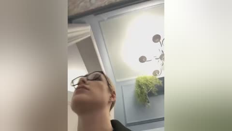 Media: A video of a young woman with fair skin, short brown hair, and glasses, looking upwards, taken from a low angle. The background features a modern room with a light-colored ceiling and a hanging fern.